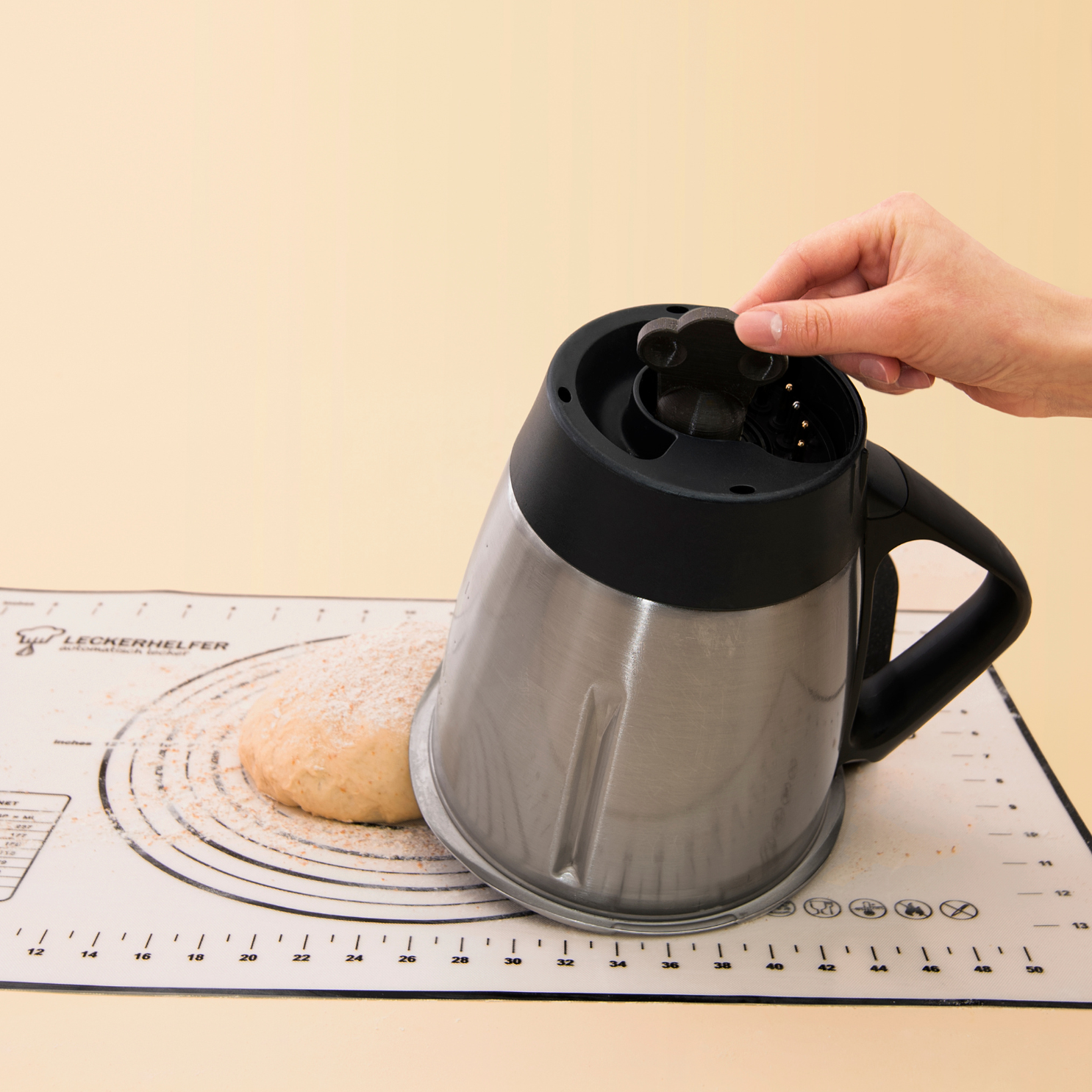 Teigdreher wird von Hand auf Schraube des umgedrehten, leicht gekippten Mixtopf eines Thermomix gesteckt. Unter dem Topf liegt ein mit Mehl bestreuter Teig auf einer Backunterlage.