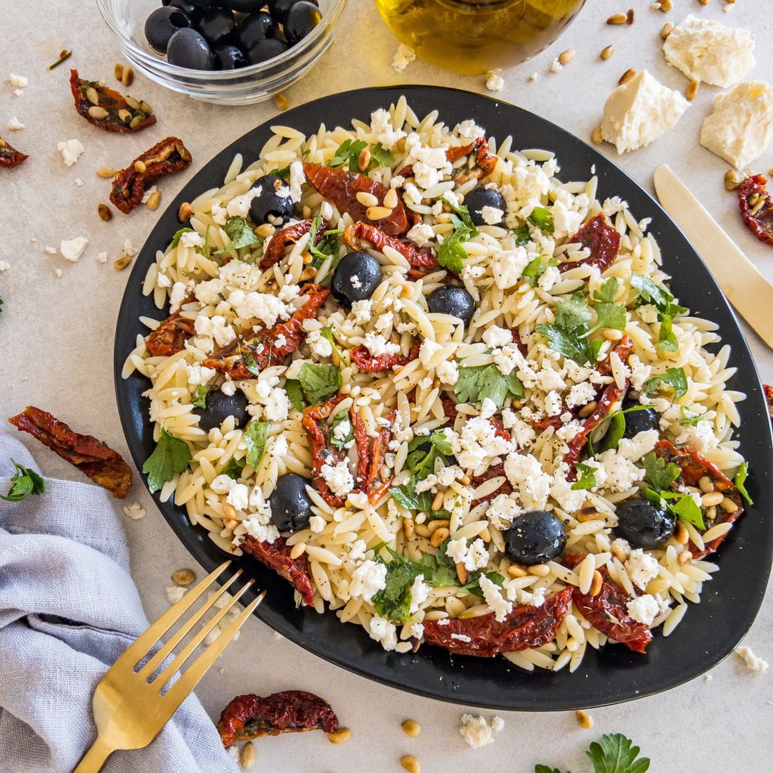 Griechischer Nudelsalat mit Feta, Oliven und getrockneten Tomaten auf einem schwarzen Teller.