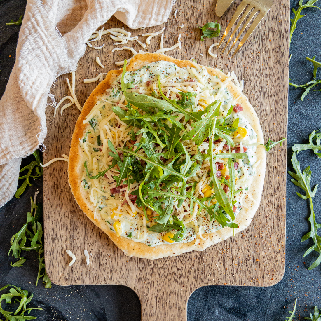 Langos garniert mit Kräutercreme, Schinkelwürfel, Streukäse und Rucola auf einem Holzbrettchen.