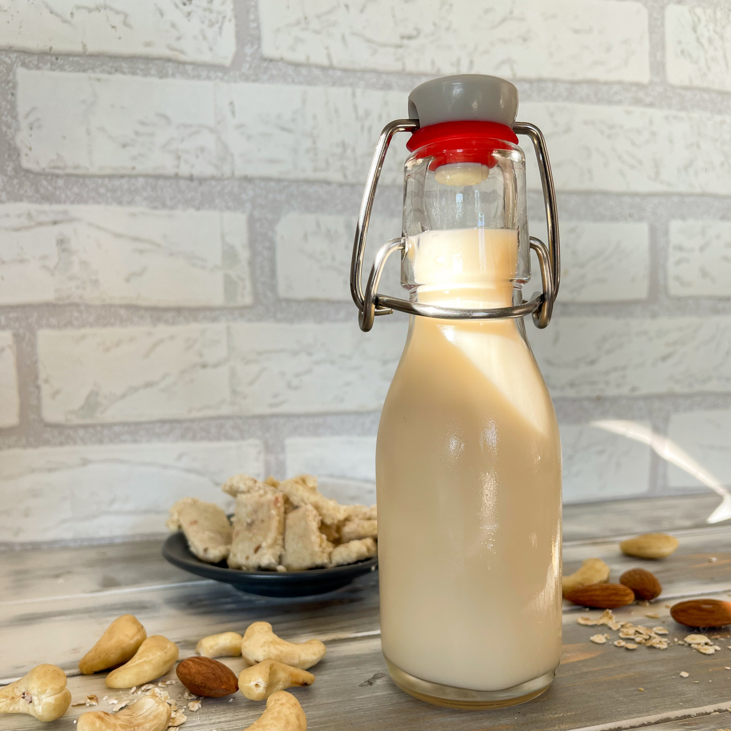 Cashewdrink in Glasflasche daneben Nüsse auf Arbeitsplatte und Müsliriegel im Hintergrund auf Teller.