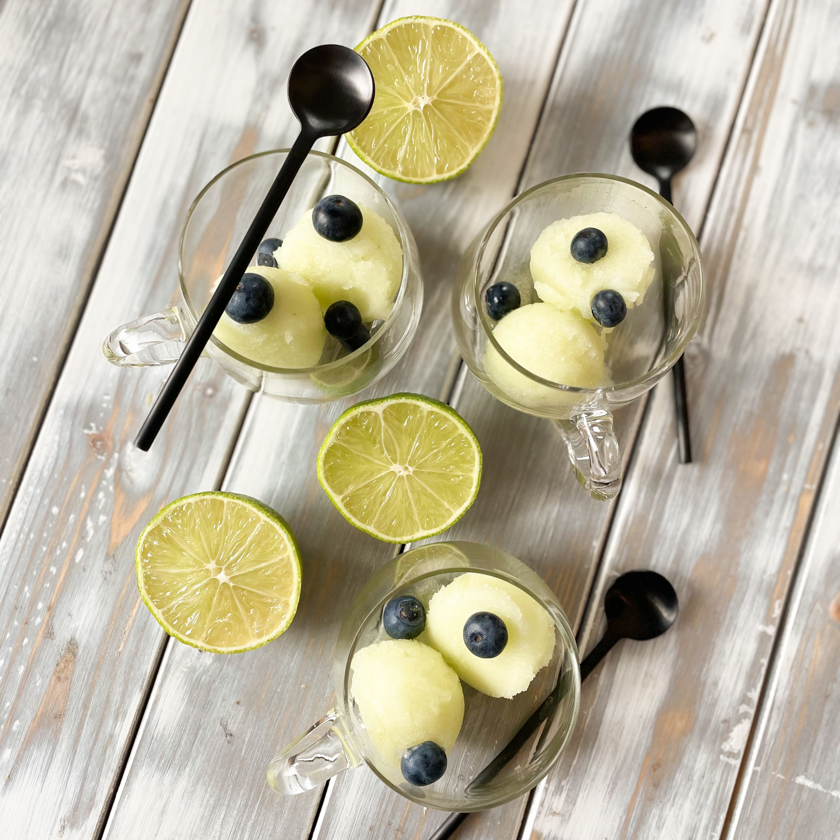 Melonen Minze Sorbet Kugeln in drei Gläsern portioniert mit frischen Blaubeeren garniert und aufgeschnittene Limetten neben den Gläsern