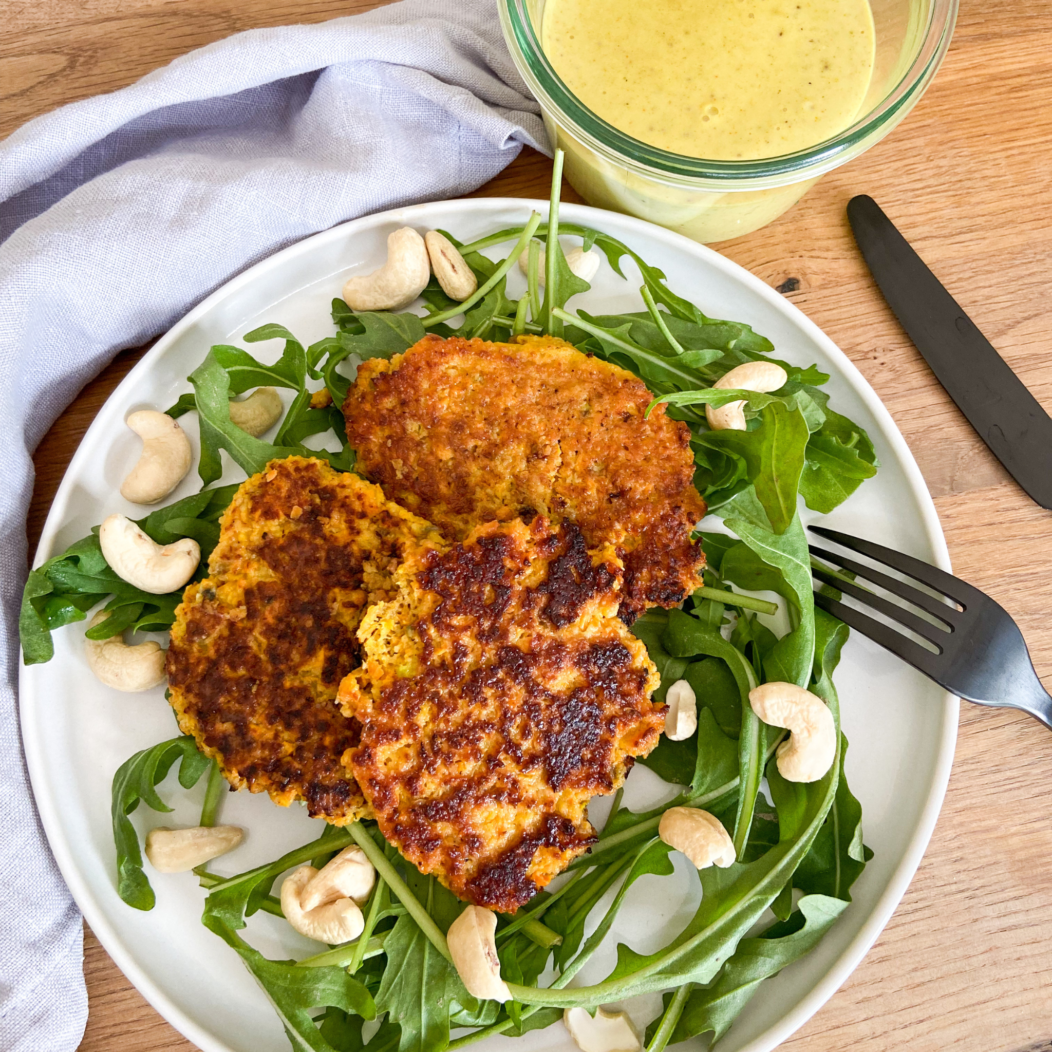 Süßkartoffelröstis auf Rucola-Salatbett daneben Cashewdip in Glasschälchen.